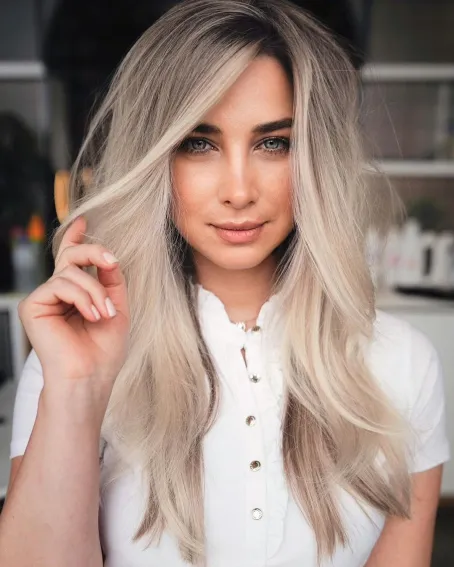 Ženska z dolgimi tankimi lasmi v pepelnato blond odtenku, postriženimi v mehke plasti in poudarjenimi z balayage prameni za več globine in volumna.